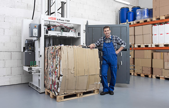 Mann steht stolz neben einer Vertikalballenpresse HSM V-Press 1160 und einem abgebundenen Ballen aus Karton